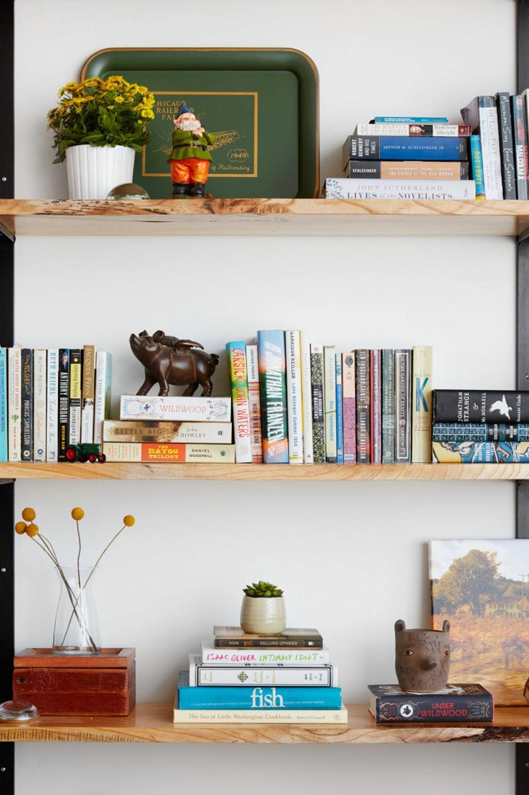 heavy-duty wall-mount bookshelves light solid wood shelving with black metal brackets. shelves have many books, vases, and figurines on them. Small gnome with green tray and paintings. Eclectic bookshelf on timeless shelving unit by Vault Furniture