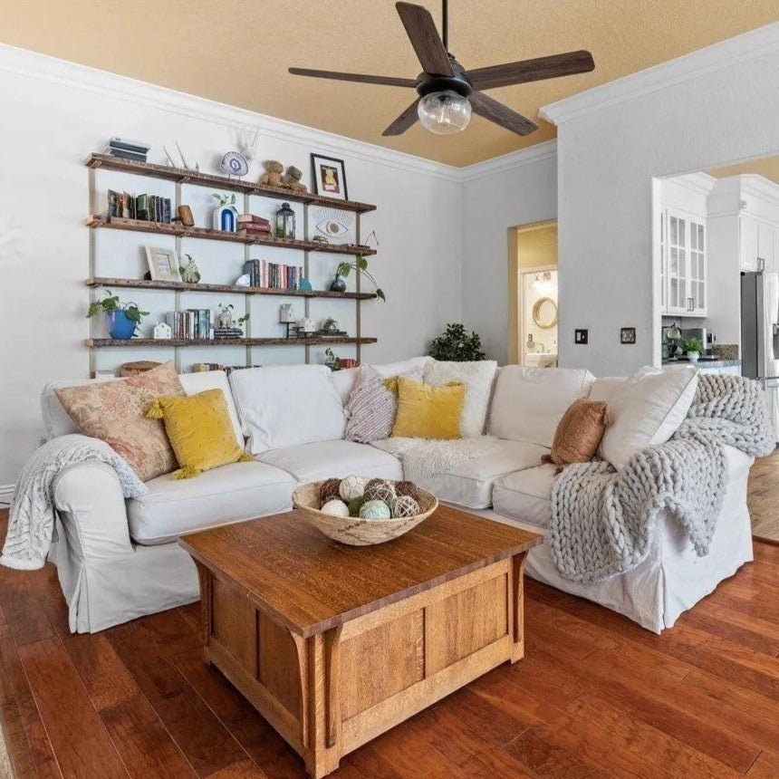 traditional living room with wall mount bookshelves, l shape white sectional and ceiling fan. large wall-mount shelving with reclaimed wood shelves and metal shelf brackets.sqaure wood coffee table on wood floors under black ceiling fan