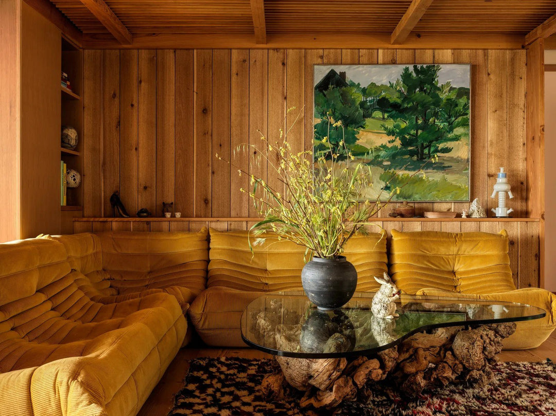cozy living room with wood panel wall and yellow velvet couch. Drift wood and glass coffee table with wood shelving 