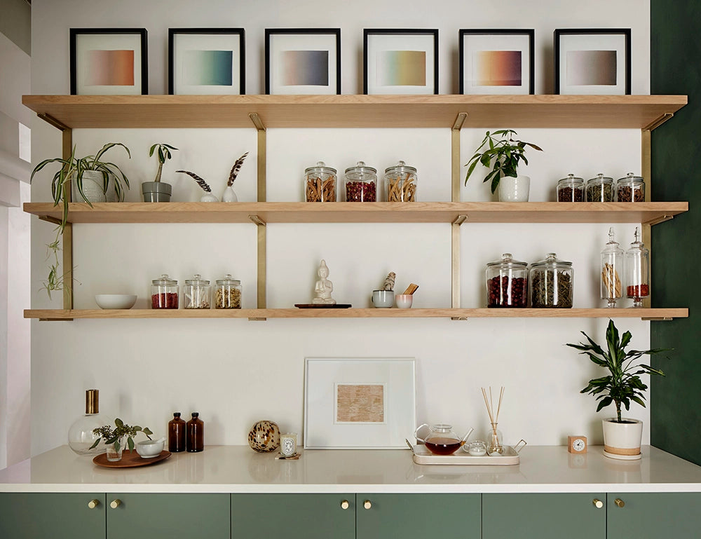 Wall-mounted wooden floating shelves with brass brackets above a kitchen counter, displaying jars, framed artwork, plants, and decorative items.