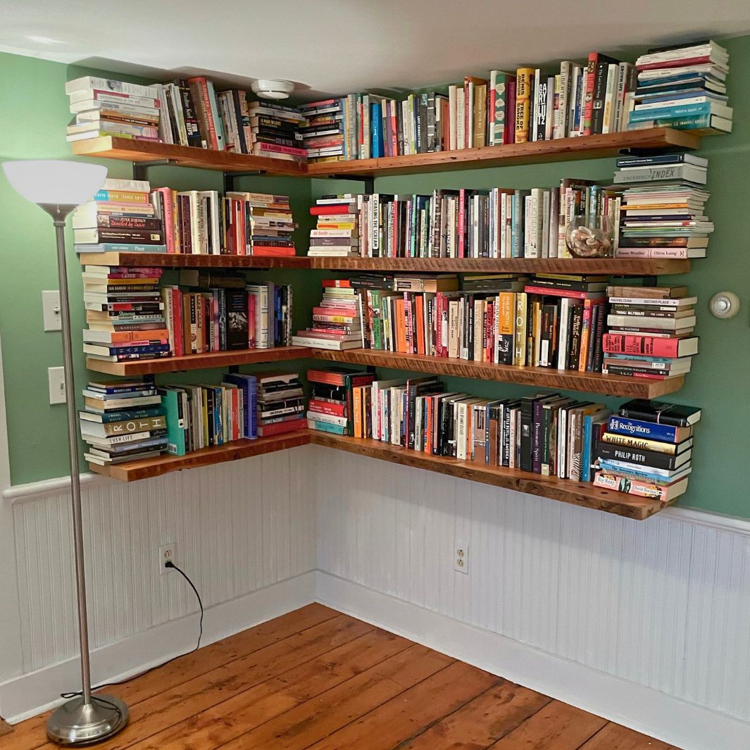 Floating bookshelves filled with books in a corner of a room. 4 wall mounted shelves with reclaimed wood and back heavy duty steel brackets. 