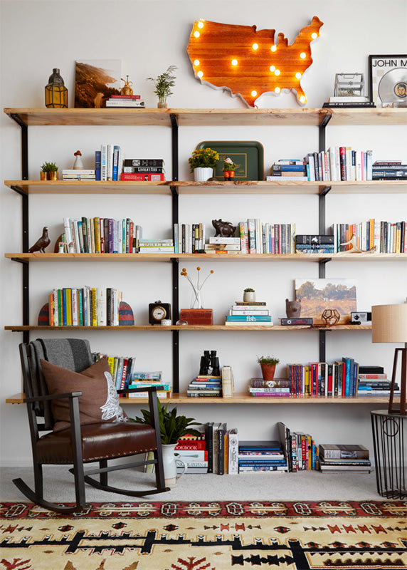 wall mounted shelving unit. tall floating blookshelves made in USA by Vault Furniture. Solid Ash wood shelving with heavy-duty black metal shelf brackets. Shelf filled with lots of books and light up america map. rocking chair sits in front of home library on top of native american rug. chicago loft style 